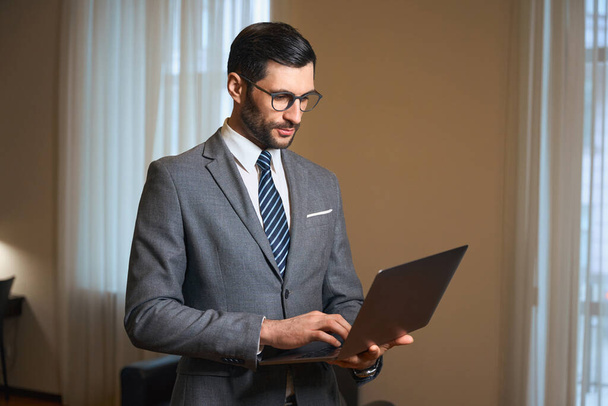 Elegant brunette is standing in a hotel room with laptop in his hands, a man is wearing stylish business suit - Foto, imagen