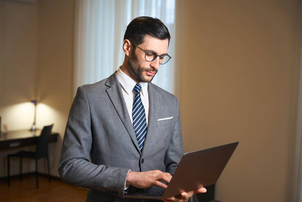 macho elegante em óculos está de pé no quarto de hotel com laptop em suas mãos, o homem está vestindo terno de negócios elegante - Foto, Imagem