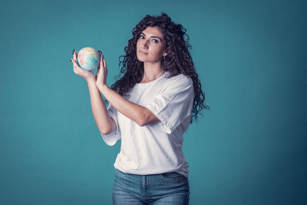 Studioaufnahme der schönen selbstbewussten kaukasischen lockigen Model Frau trägt blaue Jeans und lässiges einfaches weißes T-Shirt, isoliert über blauem Hintergrund. Schöne Brünette hält kleinen Planeten in ihren Händen - Foto, Bild