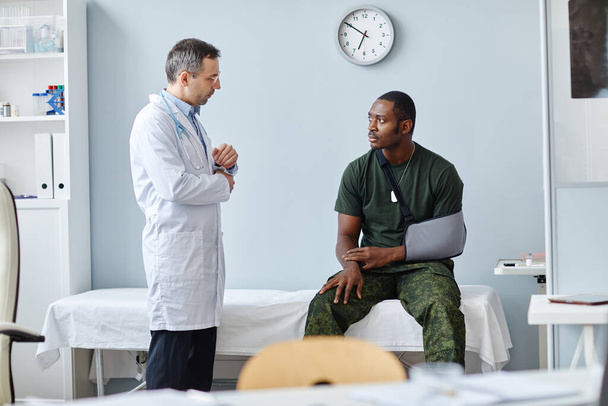 Joven oficial del ejército negro con cabestrillo de brazo sentado en el sofá médico hablando con el médico caucásico maduro - Foto, imagen
