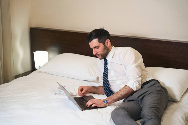 Unshaven middle-aged brunette reclining on the bed with working documents and a laptop - Φωτογραφία, εικόνα