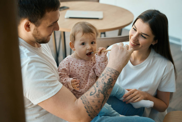 Happy dad and mom feed their baby daughter at home in kitchen, the man has a tattoo on his arm - Valokuva, kuva