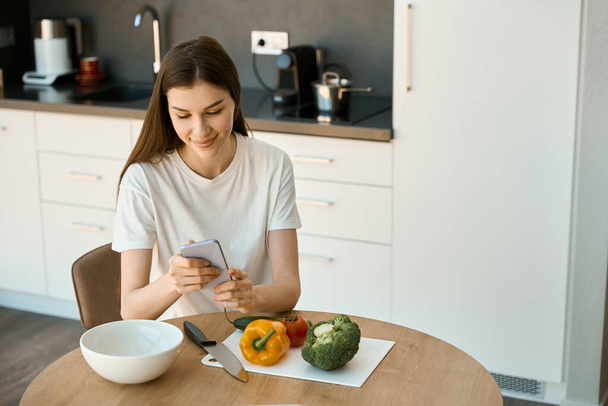 Young woman is sitting at the kitchen table with a mobile phone in her hands, vegetables are on the table - Foto, afbeelding