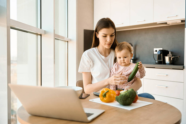 Cute young woman is babysitting a child in the kitchen, on the kitchen table there are laptops, vegetables, documents - Photo, Image