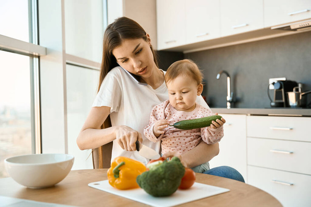 Young mother is babysitting her pretty little daughter in the kitchen, the woman is cutting vegetables - Foto, imagen