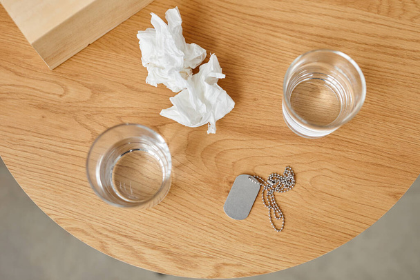 Stillleben aus gebrauchten Papierservietten, Gläsern mit Wasser und militärischem Hundeanhänger auf dem Tisch, PTBS-Behandlungskonzept - Foto, Bild