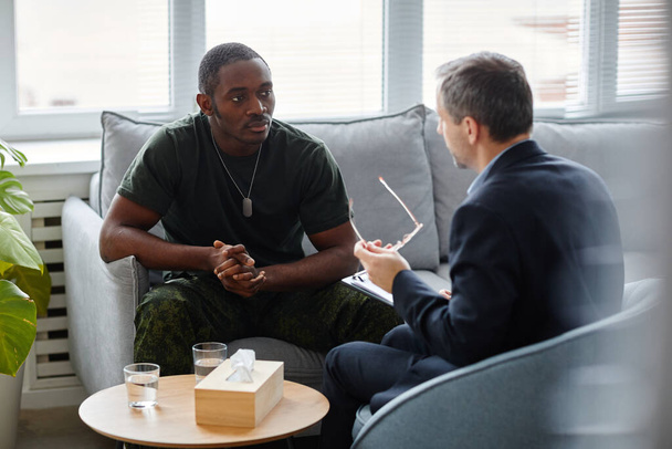 Giovane uomo afroamericano in uniforme militare seduto sul divano ad ascoltare psicoterapeuta durante la sessione di terapia - Foto, immagini
