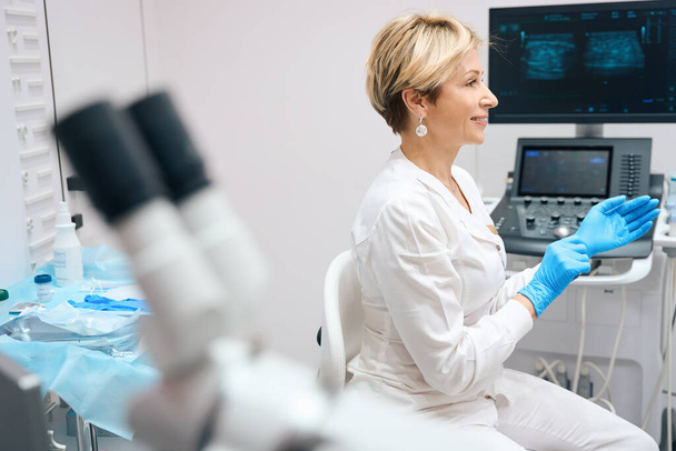 Foto einer freundlichen Gynäkologin, die medizinische Handschuhe anzieht, bevor sie die Patientin vor dem Hintergrund eines Ultraschallgeräts untersucht - Foto, Bild