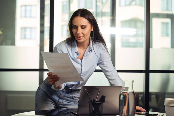 Focused corporate worker leaning on desk before laptop while looking at document in her hand - Photo, image