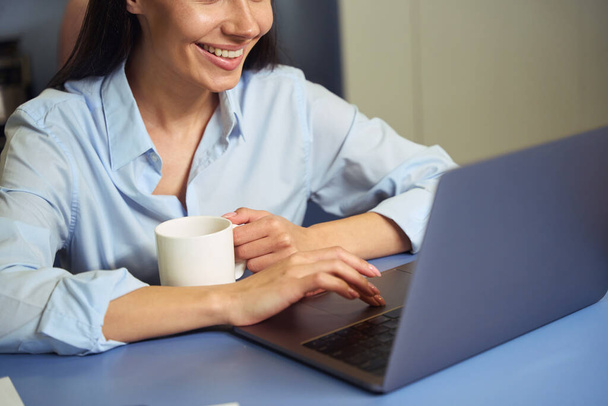 Foto recortada de la hembra sonriente con la taza en la mano sentada a la mesa haciendo clic en el touchpad portátil - Foto, imagen