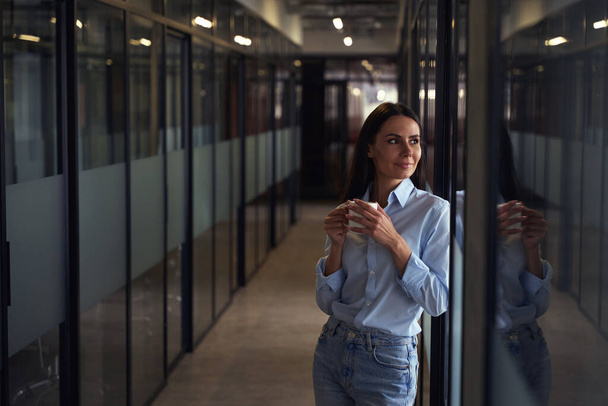 Dromerige jonge corporate werknemer met beker in haar handen leunend tegen glazen wand in gang - Foto, afbeelding