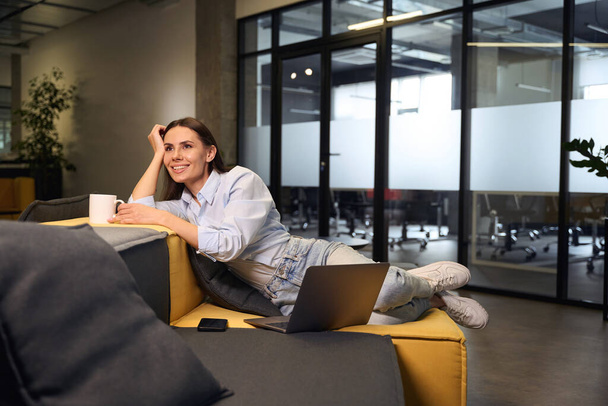 Happy kobieta pracownik biurowy z kubek i przenośny komputer siedzi na kanapie w salonie - Zdjęcie, obraz