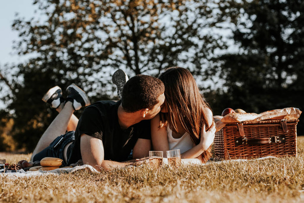 One beautiful Caucasian young couple lies on their stomachs on a bedspread with a wicker basket, fruits and gently kisses in the park against the backdrop of an old windmill, close-up side view. - Fotografie, Obrázek