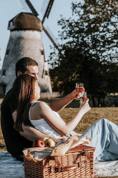 Una hermosa pareja joven caucásica sosteniendo copas de champán se sienta a medio lado en un abrazo en una colcha con una canasta de mimbre y frutas en el parque contra el telón de fondo de un viejo molino en un día soleado de verano, vista lateral de primer plano. - Foto, imagen