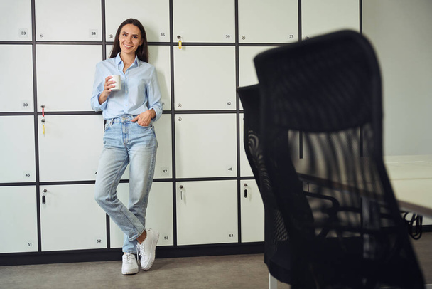 Full-size portrait of smiling young woman with mug in hand leaning against storage locker cabinet - Photo, image