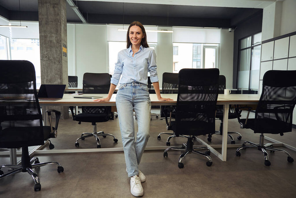 Full-length portrait of cheerful young corporate worker leaning against table in open-plan office - Foto, Imagen