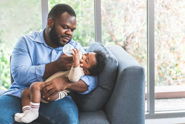Африканський батько Сидячи на дивані і годуючи молоко від пляшкового молока до його 10-місячного новонародженого сина, до сім'ї і їжі для новонародженої концепції.. - Фото, зображення