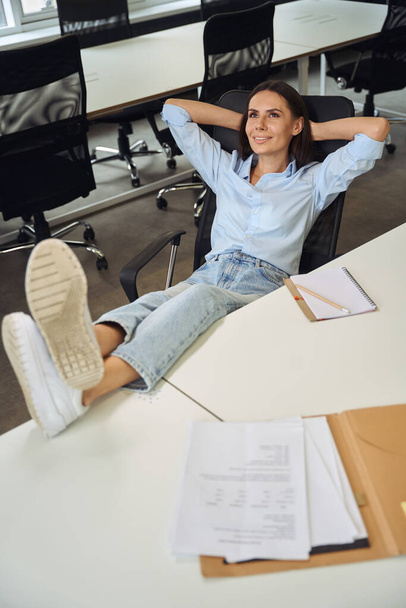 Sonriente empleada corporativa soñadora sentada en una oficina de planta abierta con los pies sobre la mesa - Foto, Imagen