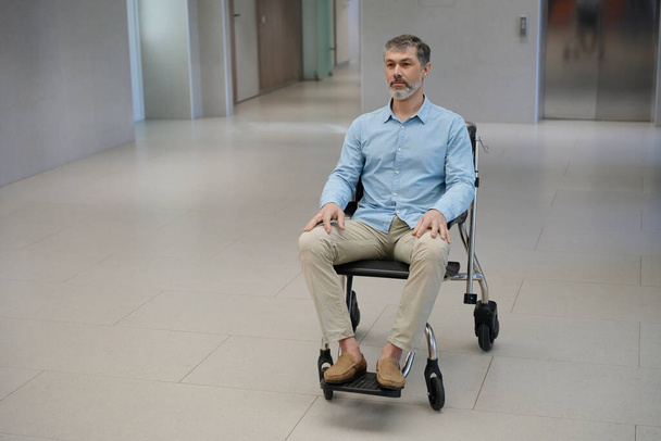 grijsharige man met een baard in een rolstoel in de gang van een moderne kliniek - Foto, afbeelding