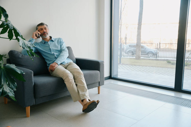 Man sitting on a black sofa near a large window and indoor plants in the corridor of the clinic - Φωτογραφία, εικόνα