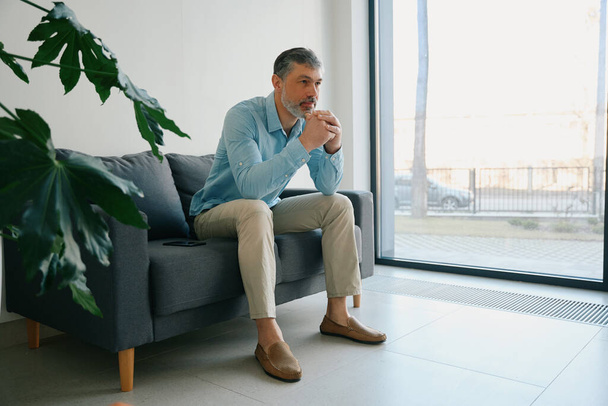 Homme avec la barbe appuyée sur ses mains est assis sur le canapé noir près de la fenêtre et des fleurs en pot dans le couloir de l'hôpital - Photo, image