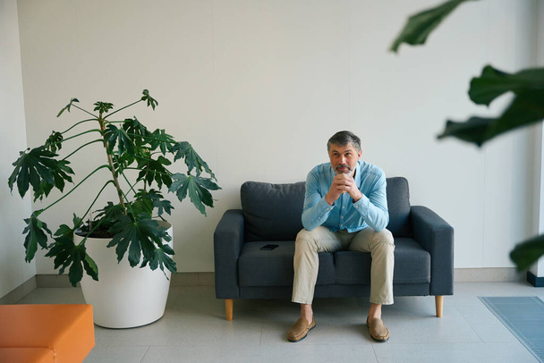 Retrato de un hombre apoyado en sus manos y sentado en un sofá rodeado de plantas de interior en un hospital - Foto, Imagen
