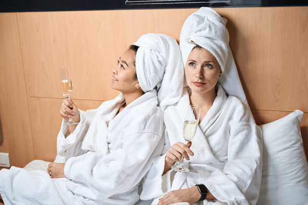 Жінки в халатах і з шампанським у ліжку. Дві подружки в білих пальто в окулярах в готелі
 - Фото, зображення