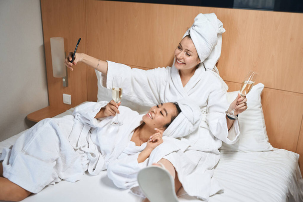Otel odasındaki mutlu bayanlar yatakta dinleniyor. Kız arkadaşlar seyahati kutlarken iyi eğlenceler. - Fotoğraf, Görsel
