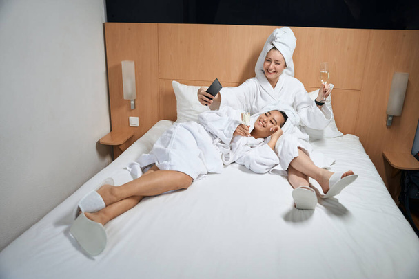 Girlfriends taking a selfie in a hotel room. Two women resting on a bed in a hotel room - Photo, image