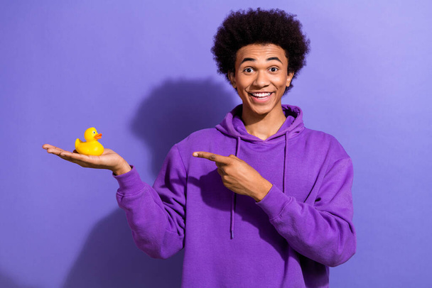 Photo of optimistic impressed guy with afro hairstyle dressed violet hoodie indicating at duck toy isolated on purple color background. - Fotoğraf, Görsel