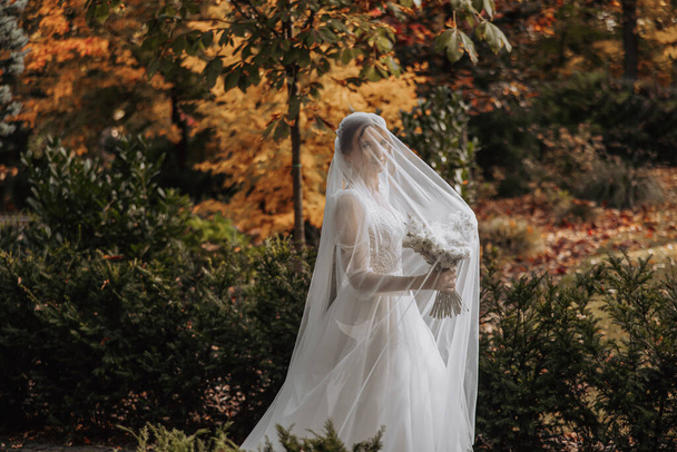 Beautiful bride with closed eyes, standing in an autumn park, holding her bouquet. Portrait of a young gorgeous bride looking into the lens. Wedding. Portrait with a veil over her face - Photo, image