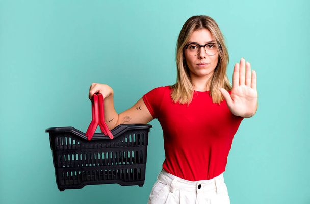 young pretty woman looking serious showing open palm making stop gesture. empty shopping basket concept - Photo, image