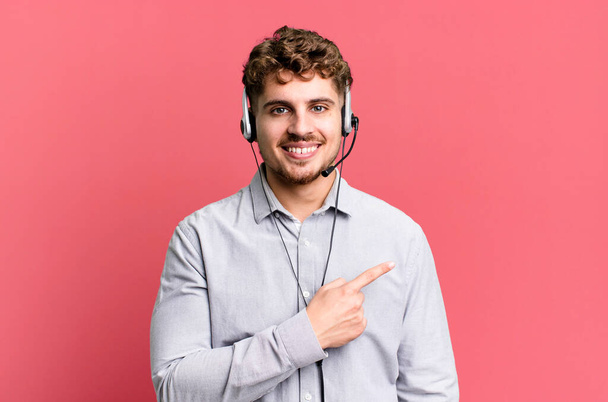 young adult caucasian man smiling cheerfully, feeling happy and pointing to the side. telemarketer agent concept - Foto, Imagen