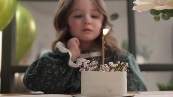 Komik küçük kız, çiçeklerle süslenmiş doğum günü pastasına yanan mum üflemeye çalışıyor. Mutlu çocuk şık bir elbise giyer ve doğum gününü dekore edilmiş oturma odasında kutlar.. - Video, Çekim