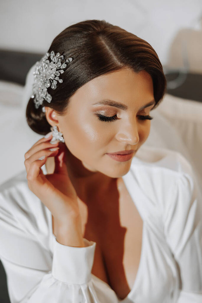Hairstyle with an elegant wedding hair accessory. The bride gently touches and shows the earrings. Gentle and stylish makeup - Fotó, kép