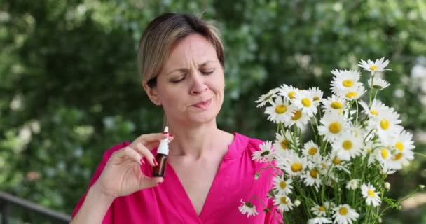 Mujer enferma e insatisfecha rocía medicina nasal caminando en floreciente jardín de primavera y sosteniendo flores de manzanilla. Tratamiento antialérgico y sintomático de la rinitis - Imágenes, Vídeo