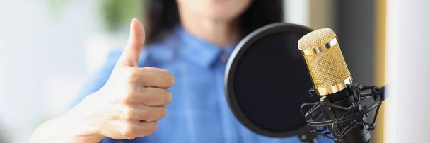 Κοντινό πλάνο της γυναίκας με ακουστικά μπροστά από το μικρόφωνο στο στούντιο που δείχνει τον αντίχειρα μέχρι την πινακίδα. Ραδιοπαρουσιαστής τραγουδιστής ή streamer έννοια - Φωτογραφία, εικόνα