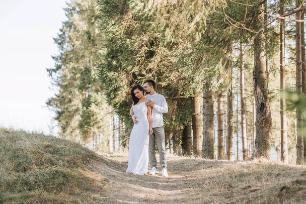 Stylish model couple in white clothes kissing at sunset in the forest. A girl hugs her husband, hair blowing in the wind. Wide angle photo - Fotó, kép
