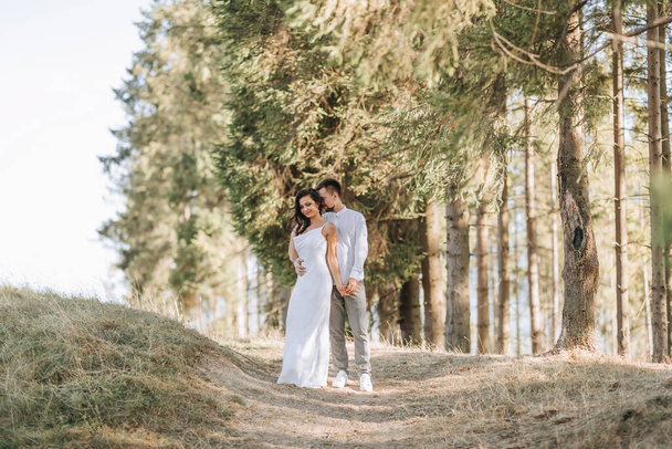 Stylish model couple in white clothes kissing at sunset in the forest. A girl hugs her husband, hair blowing in the wind. Wide angle photo - Foto, imagen