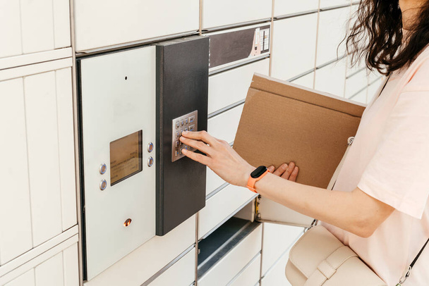A woman with a box in his hands near the self-service mail terminal. Parcel delivery machine. Person holding a cardboard box. Mail delivery and post service, online shopping, e commerce concept - Photo, Image