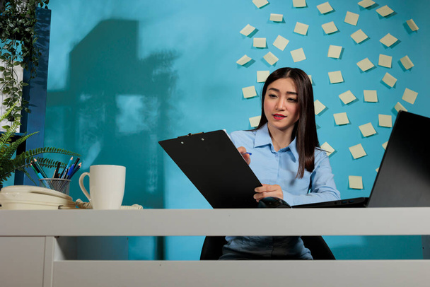 Corporate Aziatische werknemer met behulp van laptop om een online onderzoek te doen en notities te maken op klembord. Zakenvrouw die aan het bureau zit en zich concentreert op het maken van aantekeningen door het formulier in te vullen. - Foto, afbeelding
