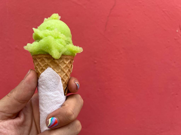 Mužova ruka drží zelenou, citrónovou kopeček zmrzliny v kuželu u červené stěny. Člověk má manikúru na nehty. - Fotografie, Obrázek
