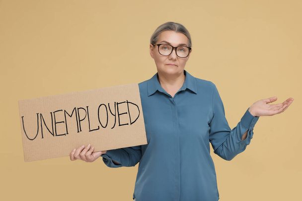Confused senior woman holding cardboard sign with word Unemployed on beige background - Foto, Imagen