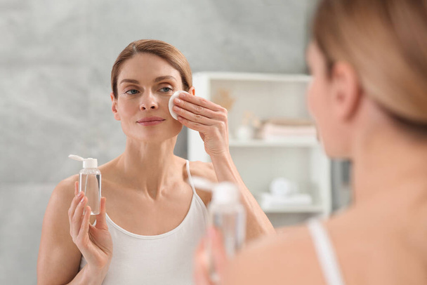 Beautiful woman removing makeup with cotton pad near mirror indoors - Foto, Imagem