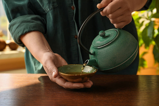 Woman pouring freshly brewed tea from teapot into cup at wooden table indoors, closeup. Traditional ceremony - Foto, Bild