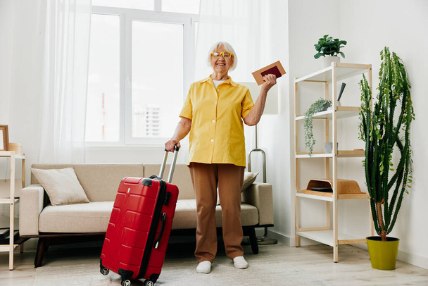 Szczęśliwa seniorka z paszportem i biletem na podróż spakowała czerwoną walizkę, wakacje i opiekę zdrowotną. Uśmiechnięta staruszka radośnie stoi w domu przed podróżą. Wysokiej jakości zdjęcie - Zdjęcie, obraz