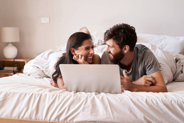Bett, Laptop und glückliches Paar entspannen sich, lächeln und freuen sich auf Webinfos, Morgenblog oder Website-Benachrichtigungen. Schlafzimmer zu Hause, Blickkontakt und Menschen, die online einkaufen oder im Internet suchen. - Foto, Bild