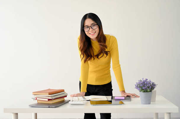 Een aantrekkelijke en gelukkige jonge Aziatische studente of kantoormedewerker in bril en casual kleding buigt voorover aan tafel en glimlacht naar de camera. geïsoleerd op een witte achtergrond - Foto, afbeelding