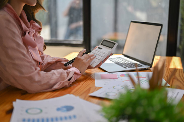 Imagen recortada de una hermosa mujer de negocios asiática o contable en un vestido rosa usando una calculadora y analizando datos financieros en su escritorio. - Foto, Imagen
