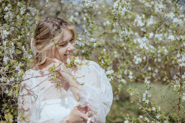 Smiling young bride enjoying nature while standing close to sakura branches. Wonderful lady in wedding dress surround by white cherry flowers, spending time outdoor on sunny summer day. - Foto, Imagem